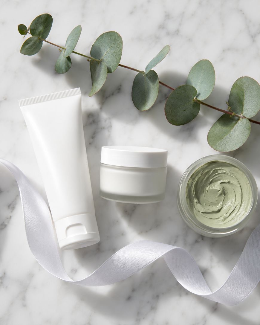 A white tube, a small white jar, and an open jar with green cream sit on a marble surface with a white ribbon and a eucalyptus branch.