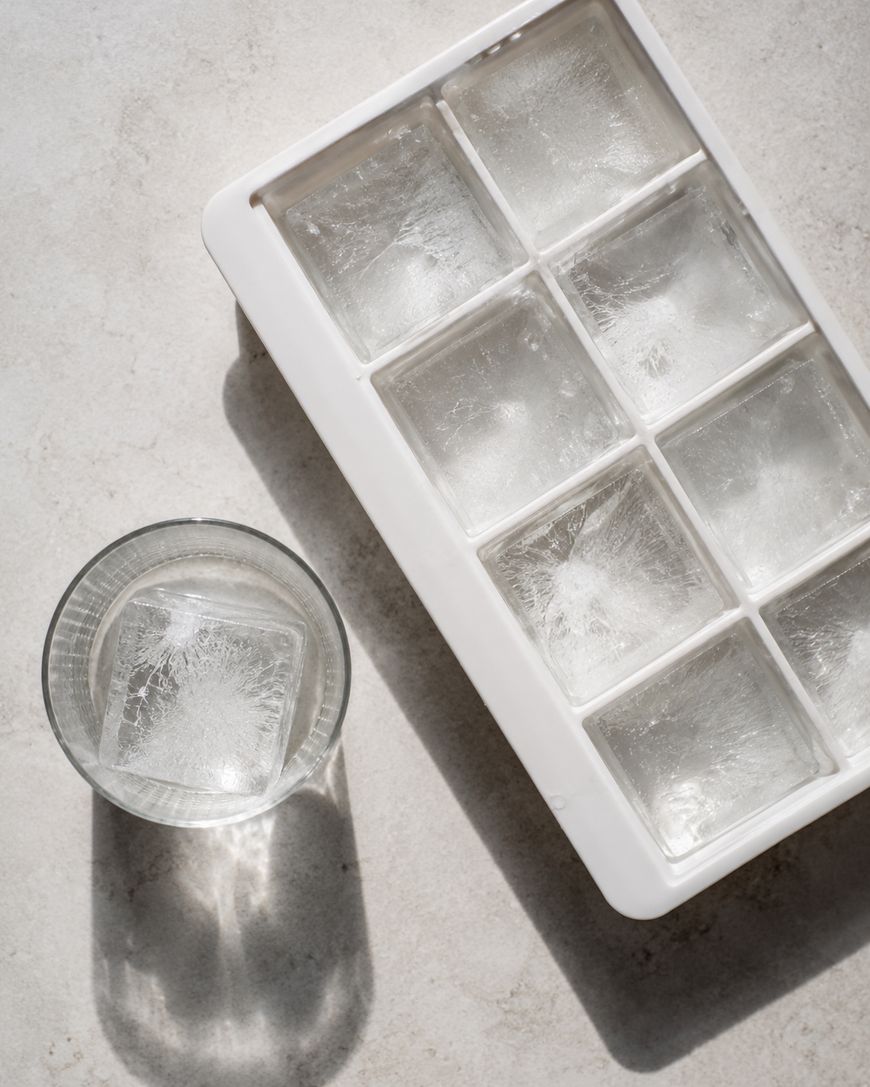 A white ice cube tray with large square ice cubes sits beside a glass of water containing one ice cube, all on a light-colored surface.