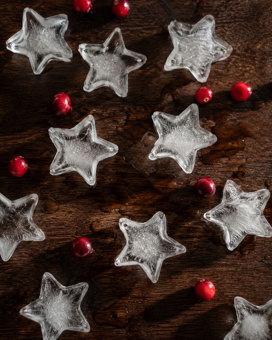 Star-shaped ice cubes scattered on a dark wooden surface with several small red cranberries placed in between the ice cubes.