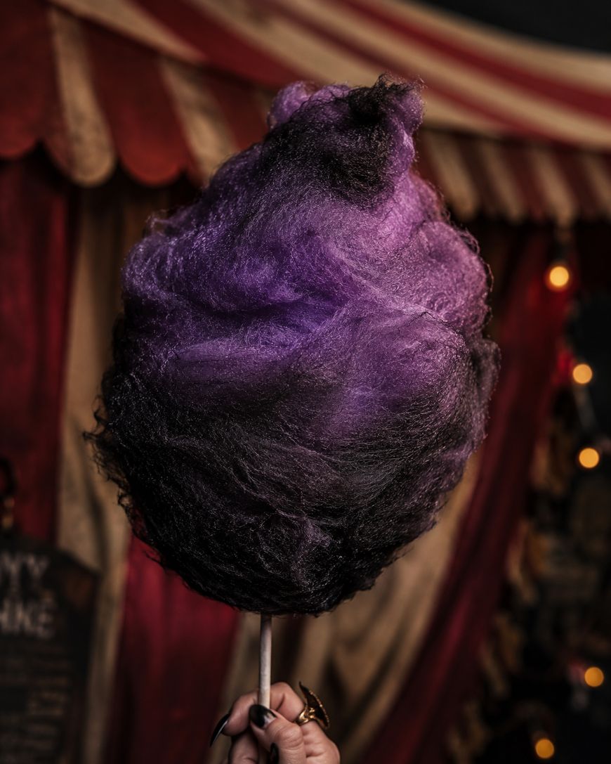 A hand holds a large stick of purple and black cotton candy in front of a circus tent with red and white stripes and dim lights in the background.