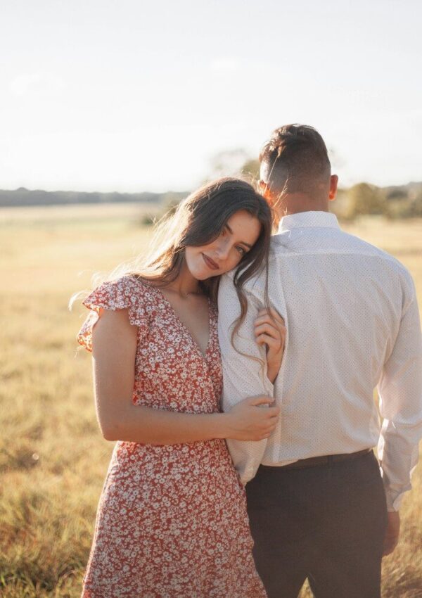 A woman in a red floral dress leans her head on the shoulder of a man in a white shirt, standing together in a sunlit field.