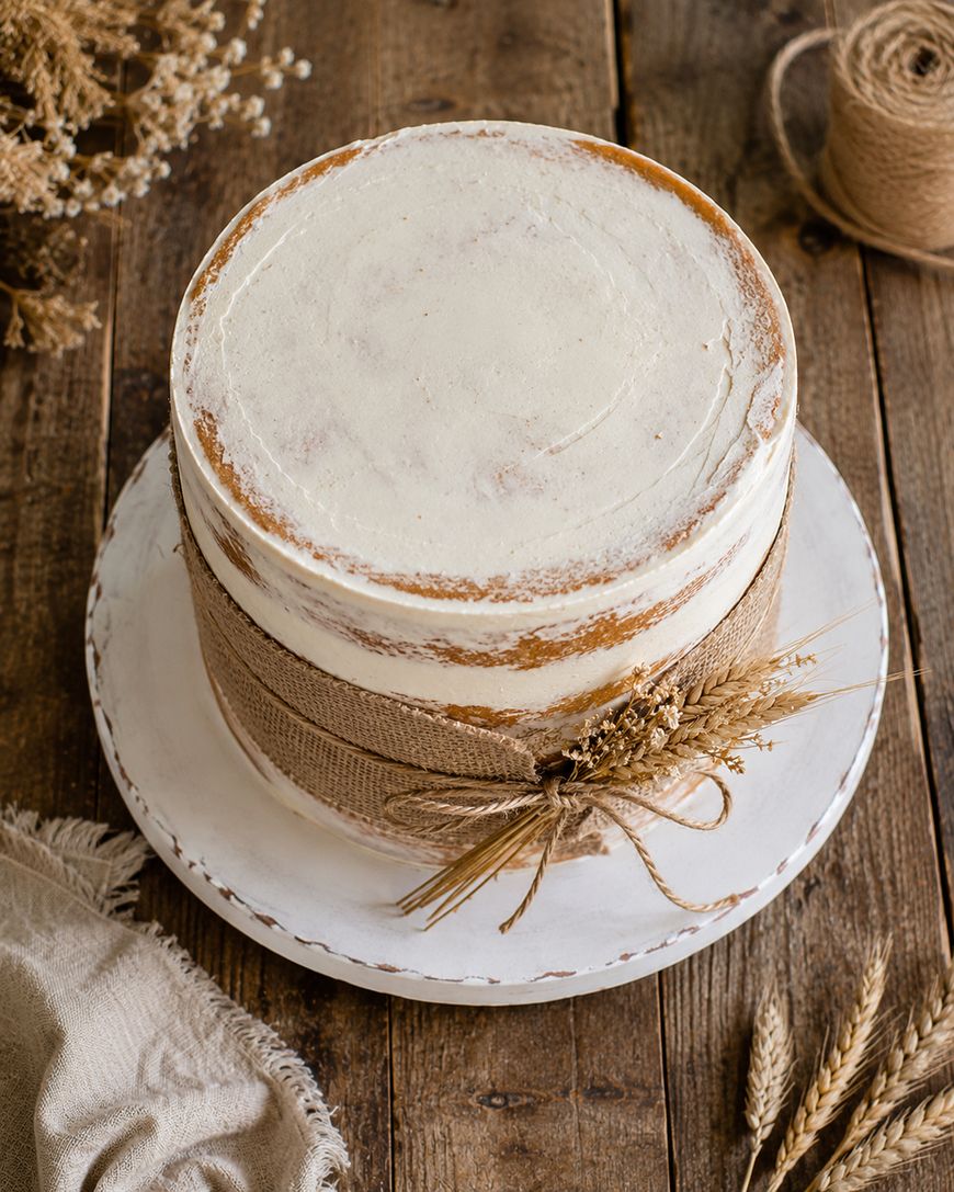 A rustic, semi-naked cake with minimal white frosting, wrapped in burlap and twine, decorated with dried wheat, sits on a white cake stand atop a wooden table.