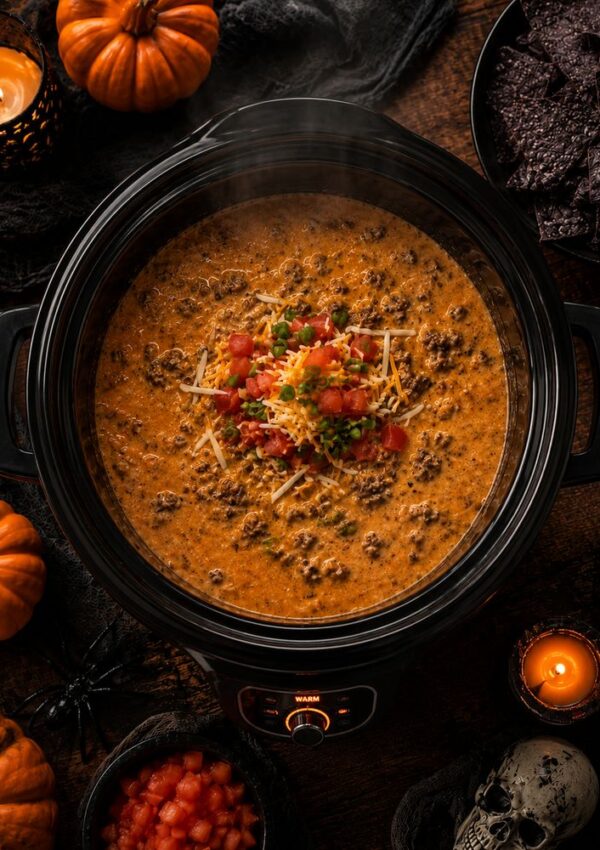 A crockpot filled with cheesy taco dip, topped with diced tomatoes and shredded cheese, surrounded by pumpkins, candles, black chips, and Halloween decorations.