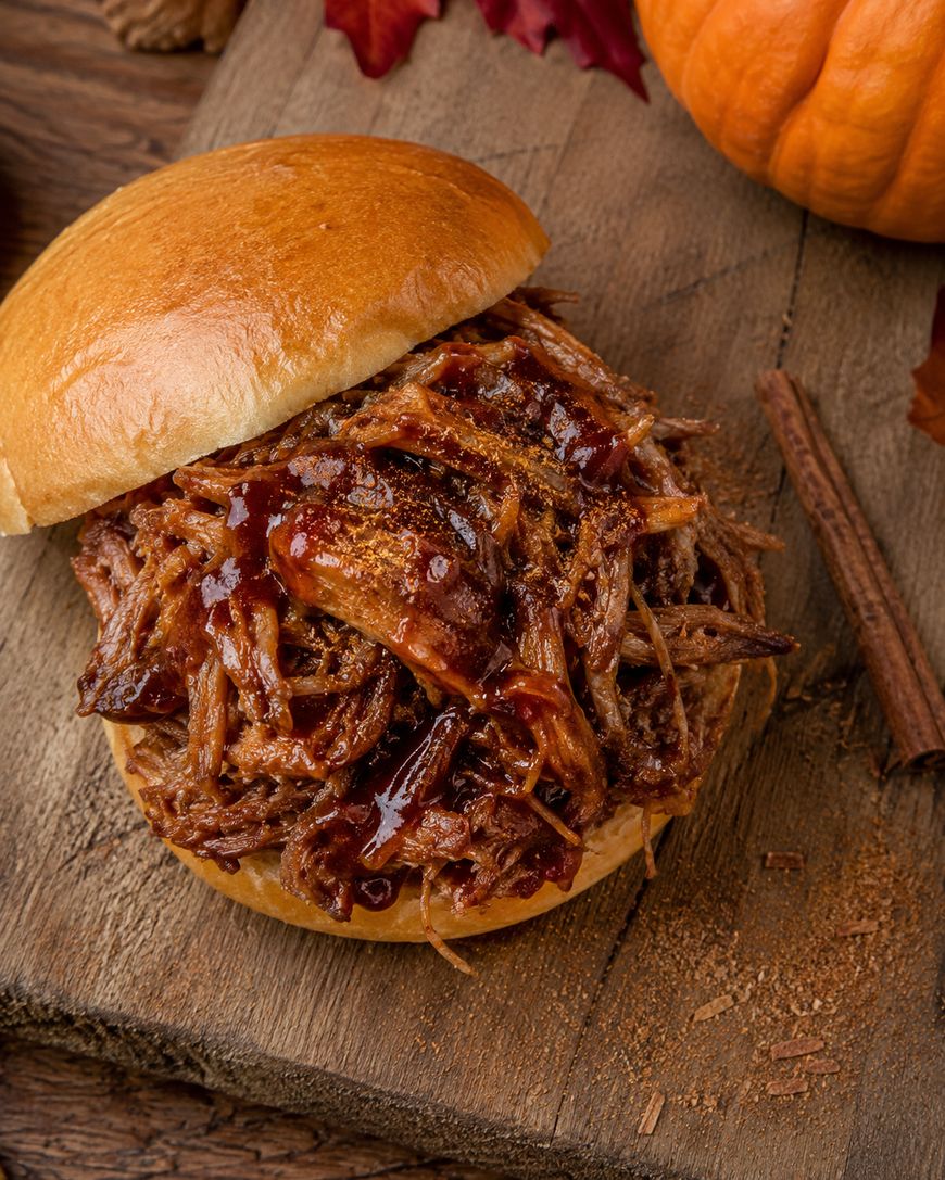 A pulled pork sandwich with barbecue sauce on a bun sits on a wooden surface, surrounded by a pumpkin, cinnamon stick, and autumn leaves.