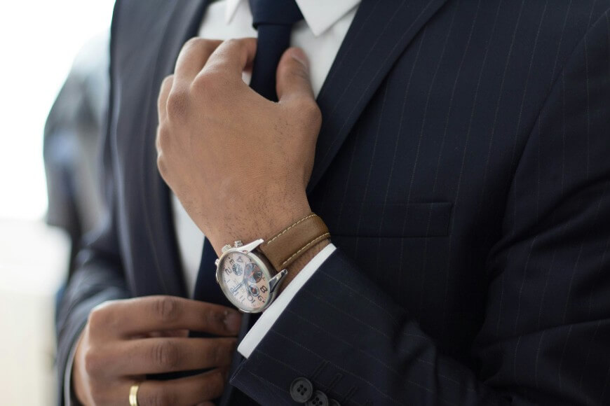 A person in a pinstripe suit adjusts a dark tie with one hand, wearing a watch on the wrist. Only the torso and hands are visible, not the face.