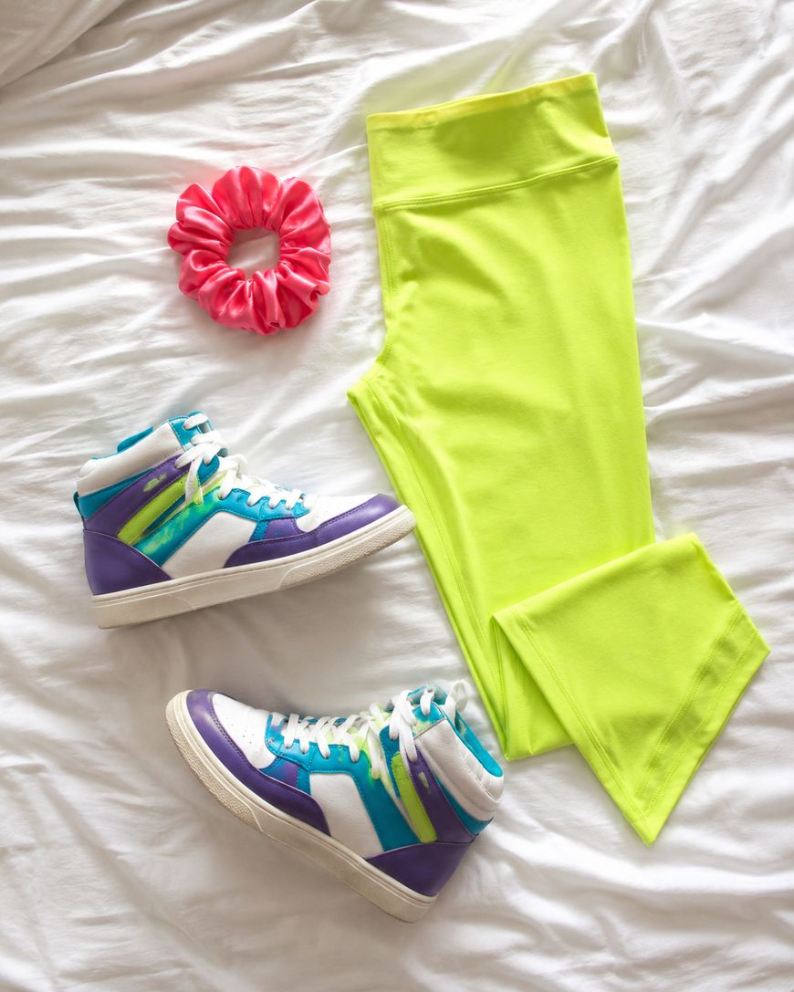 Neon yellow leggings, a pair of purple, teal, and white high-top sneakers, and a pink scrunchie are arranged on a white bedsheet.