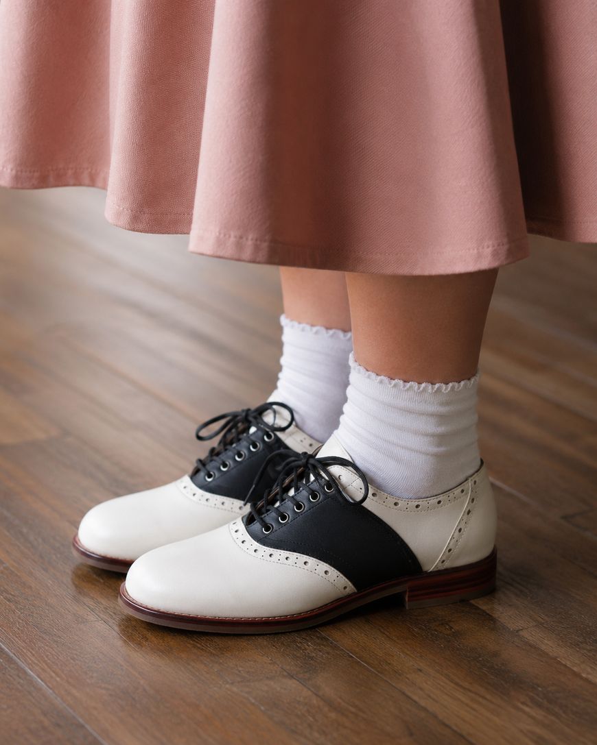 A person wearing a pink skirt, white frilly socks, and black-and-white saddle shoes stands on a wooden floor.