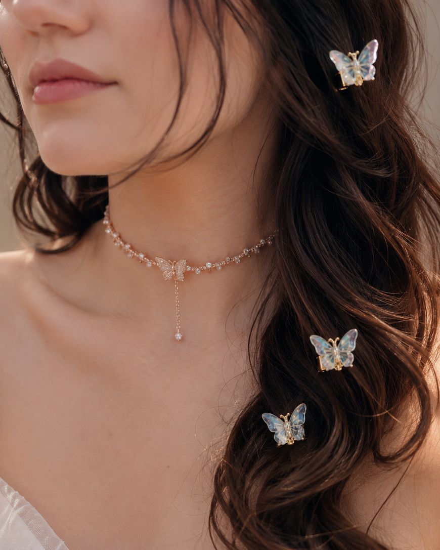 A woman with long, wavy brown hair wears a delicate butterfly necklace and pearl butterfly hair clips. She has light skin and wears a soft, off-the-shoulder top.