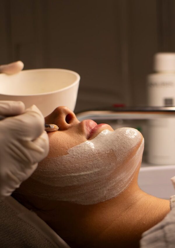 A person reclines as someone wearing gloves applies a white facial mask with a brush—an experience reminiscent of both at-home beauty routines and professional beauty treatments. A bowl is held nearby and skincare products are visible in the blurred background.