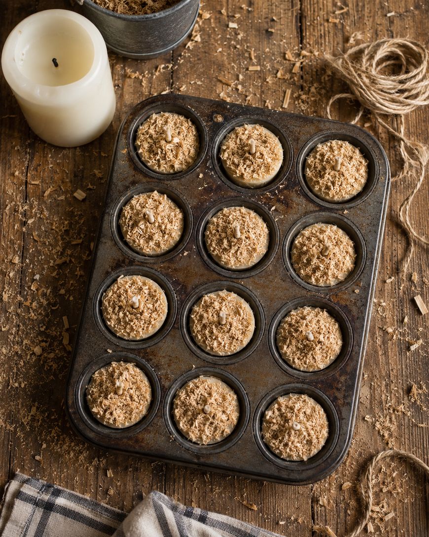 A muffin tin filled with twelve unbaked, crumbly oat muffins sits on a rustic wooden table, surrounded by a white candle, string, a bowl of oats, and a plaid cloth.