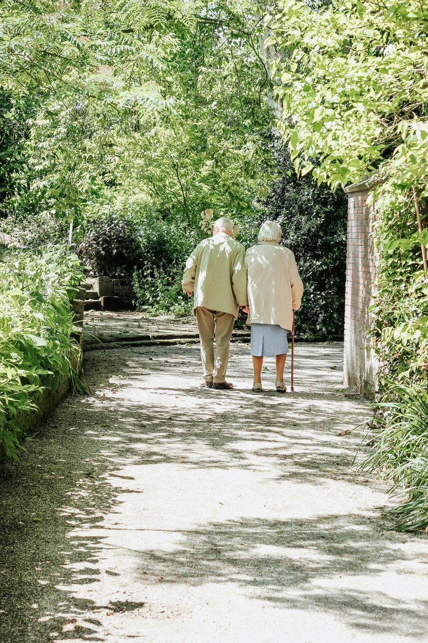 An elderly couple walks arm in arm down a sunlit garden path surrounded by lush greenery. The woman uses a cane, and both wear light-colored coats, enjoying a peaceful stroll together—an everyday moment cherished by many London families who value the comfort of care at home.