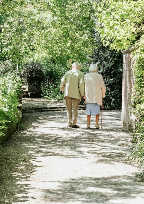 Two elderly people walk arm in arm down a sunlit garden path, surrounded by lush green plants. One uses a cane for support, enjoying the tranquillity and independence made possible through attentive care at home.