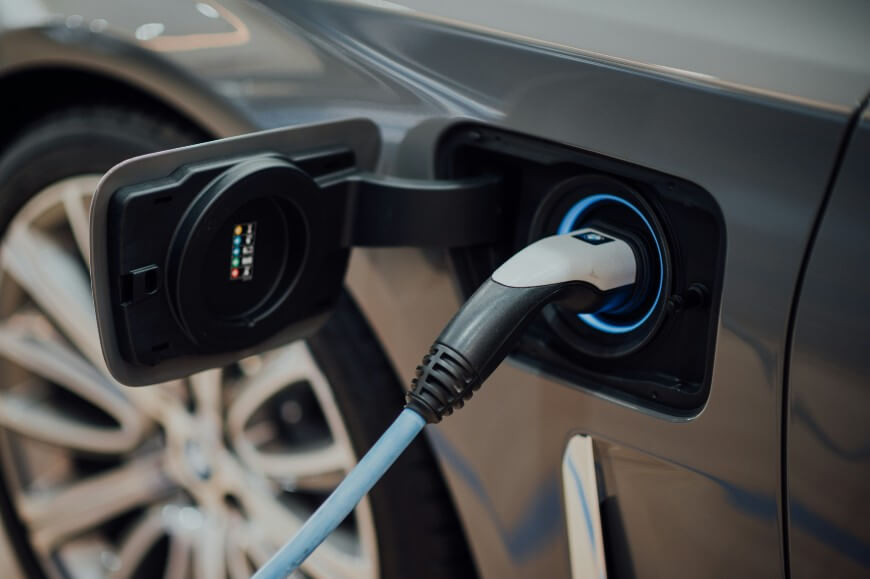 A close-up view of an electric vehicle being charged, showing a charging cable plugged into the car’s charging port with the vehicle’s wheel visible in the background.