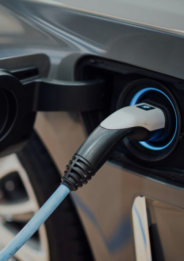 A close-up view of an electric vehicle being charged, showing the charging plug connected to the cars charging port. The car appears to be grey and the charging cable is blue.
