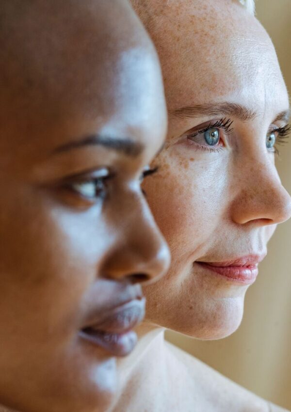 Close-up of two womens faces in profile, one with dark skin and the other with light skin, both looking ahead with calm, thoughtful expressions against a neutral background.
