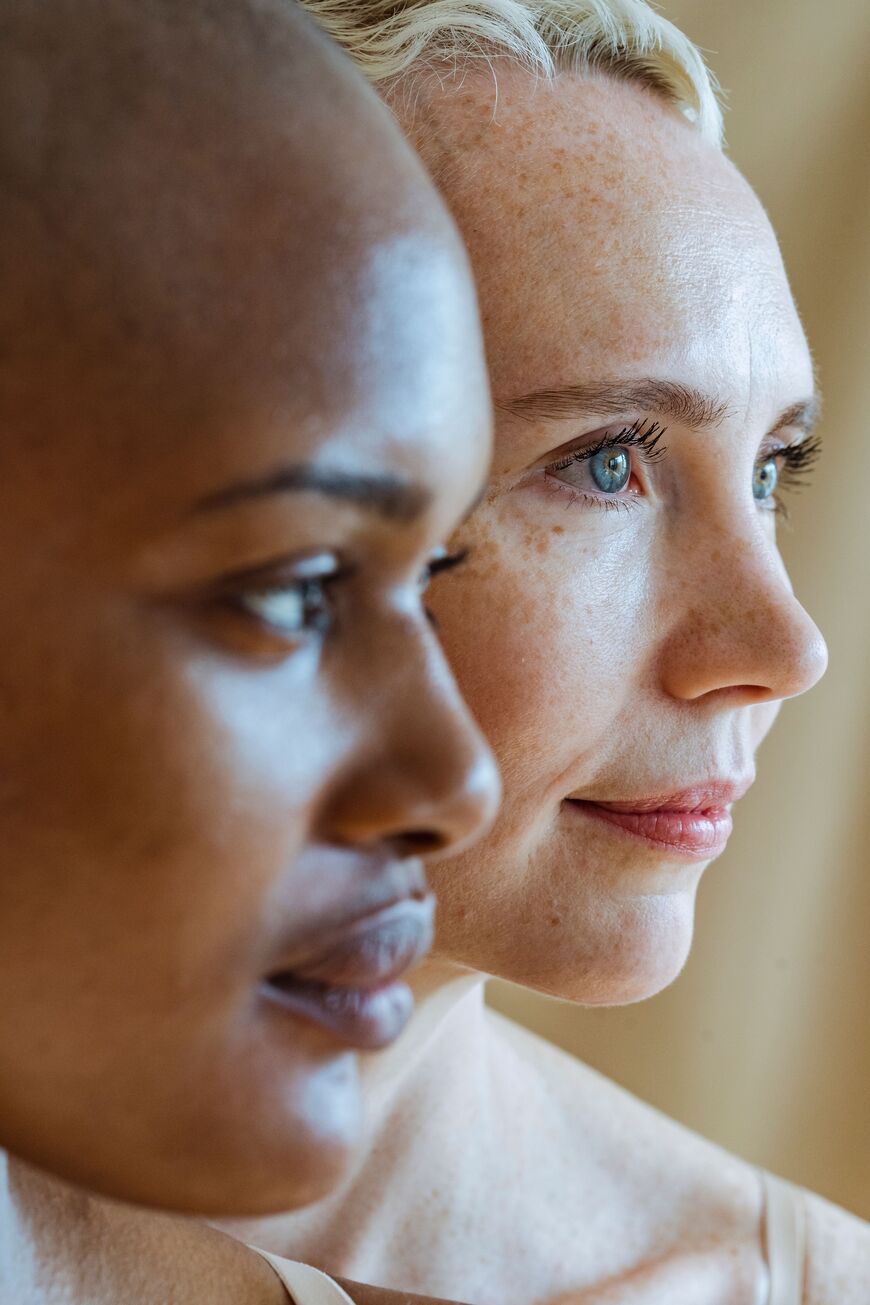 Two women stand side by side in close-up, one with dark skin and a shaved head, the other with light skin, blue eyes, and blonde hair, both looking thoughtfully into the distance.
