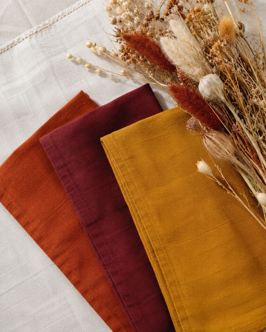 Three folded cloth napkins in rust, burgundy, and mustard yellow, next to a bundle of dried grasses and flowers, all on a white linen surface.
