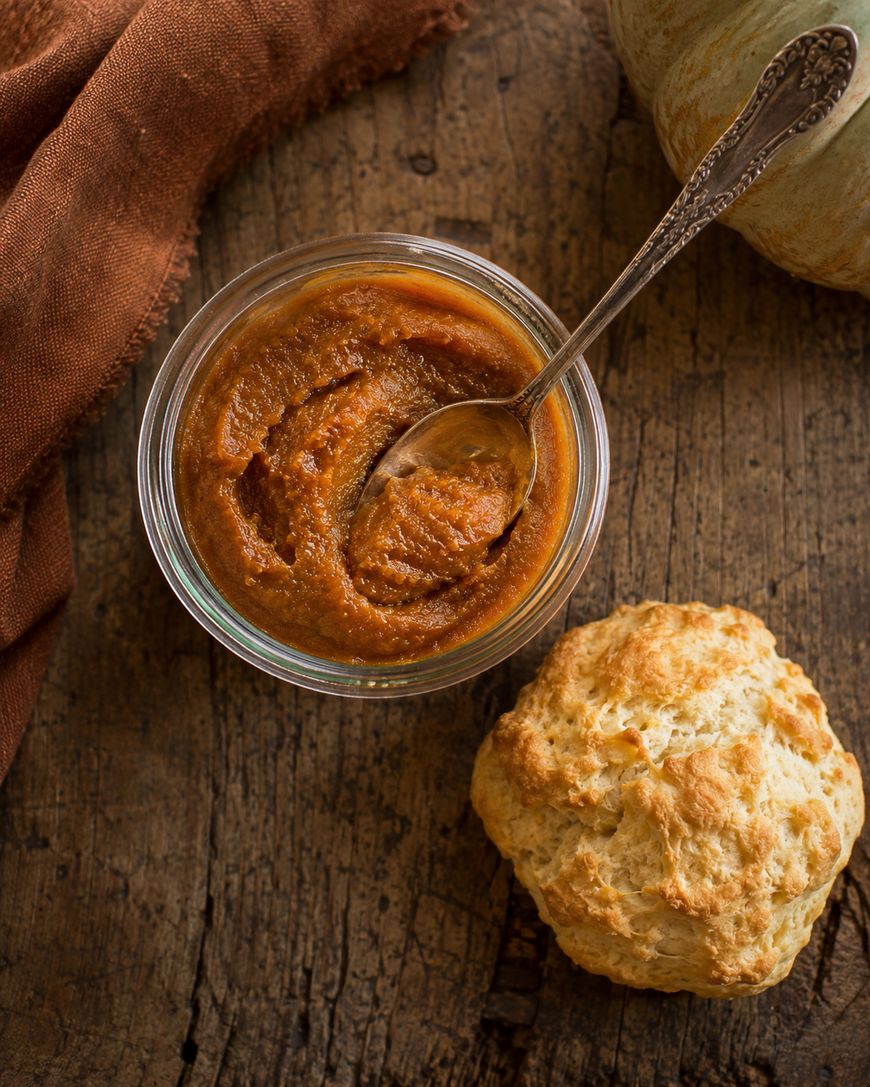 A glass bowl of pumpkin butter with a spoon, next to a golden biscuit on a rustic wooden table and a brown cloth.