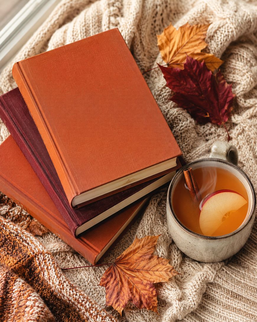 A closed book with a romantic cover showing a silhouetted couple is on a wooden table surrounded by autumn leaves, a pumpkin, cinnamon sticks, and plates with food.