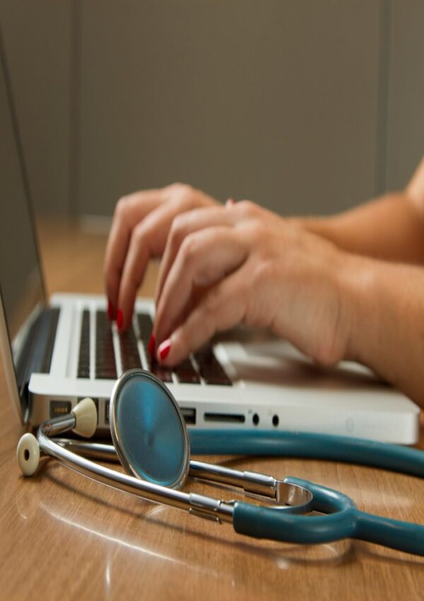 Hands typing on a laptop keyboard with a stethoscope lying on the wooden table nearby, suggesting a medical or healthcare setting.