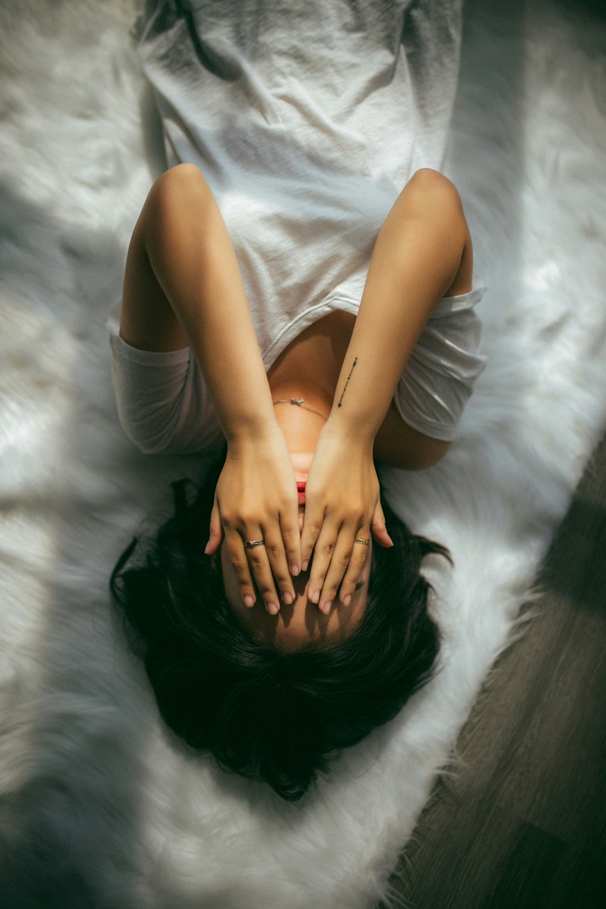 A person in a white shirt lies on a fluffy white rug, covering their face with both hands. Sunlight casts gentle shadows on the scene.