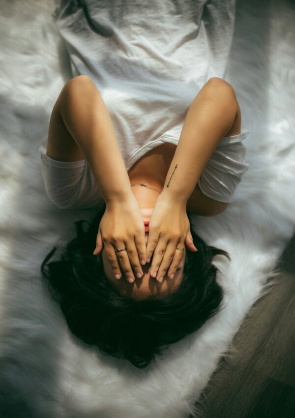 A person in a white shirt lies on a fluffy white rug, covering their face with both hands. Light and shadow fall across their body, creating a soft, serene atmosphere.