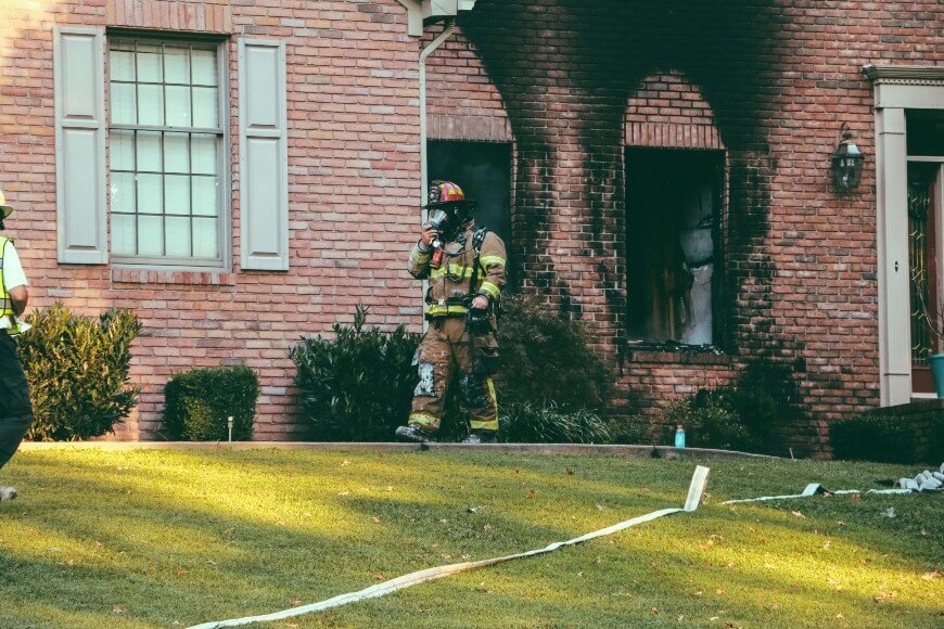 A firefighter walks outside a brick house with burned windows and blackened walls after a fire. The lawn has hoses, and another person is partly visible on the left.