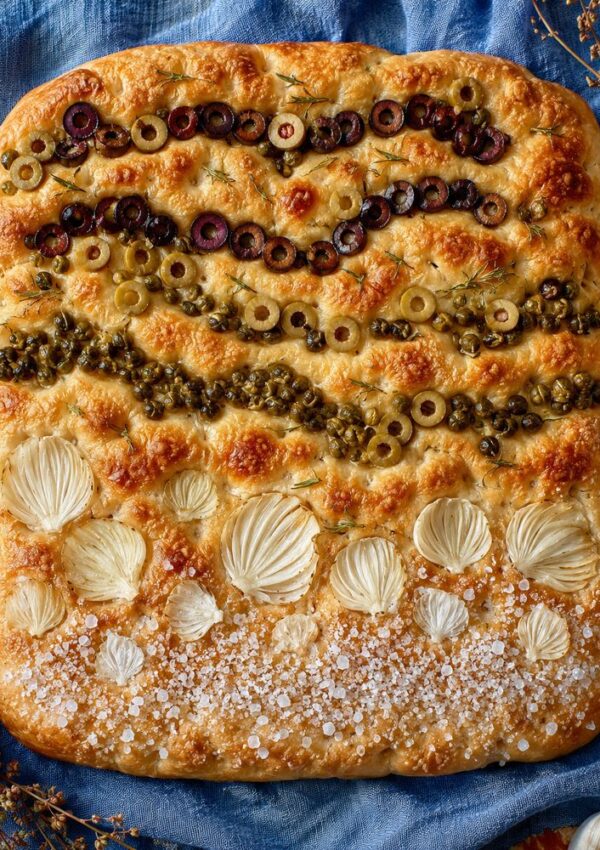 A square focaccia decorated with rows of sliced black and green olives, capers, onion petals, and coarse salt showcases stunning Focaccia bread art on a golden-brown crust, set against a blue cloth background. This is one of the most inspiring bread art ideas for anyone looking to elevate their focaccia recipes.