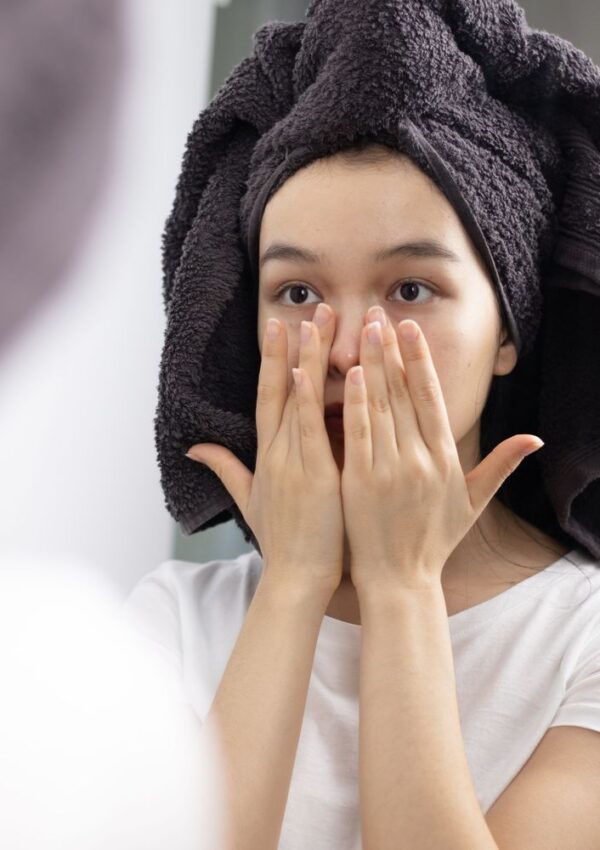 A person with a dark towel wrapped around their head touches their face gently while looking in a mirror, wearing a white shirt, as if admiring their glowing skin after following the best beauty tips.