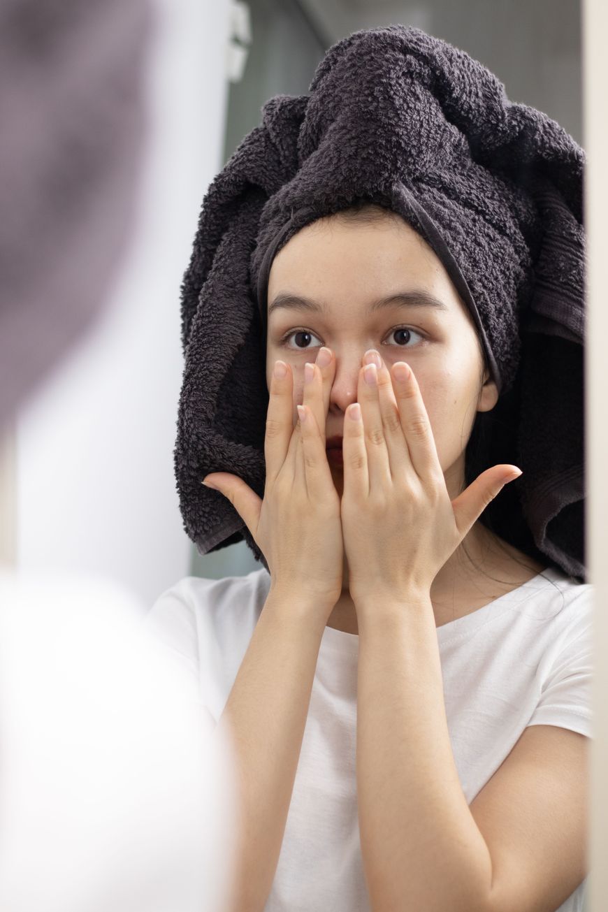 A woman with a towel wrapped around her head touches her face with both hands while looking at herself in a mirror, reflecting on the best beauty tips for achieving glowing skin.