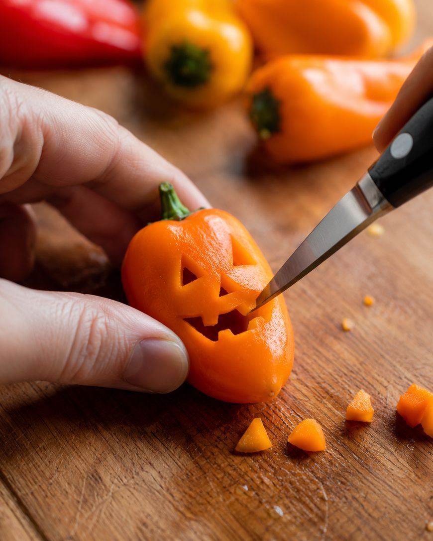 A hand carves a jack-o-lantern face into a small orange pepper with a knife on a wooden surface. Other whole peppers and small orange pieces are nearby.