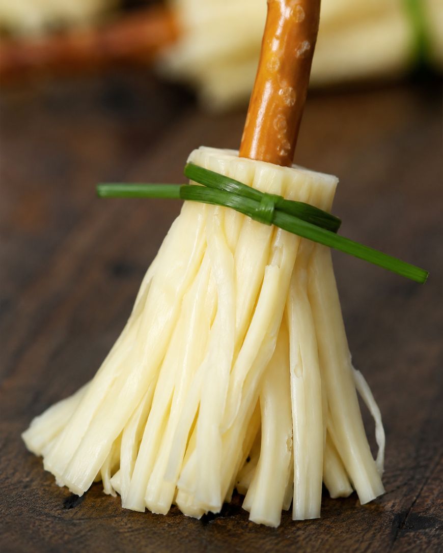 A snack shaped like a broom: strands of string cheese form the bristles, tied with a green chive, and a pretzel stick serves as the handle, all placed on a wooden surface.