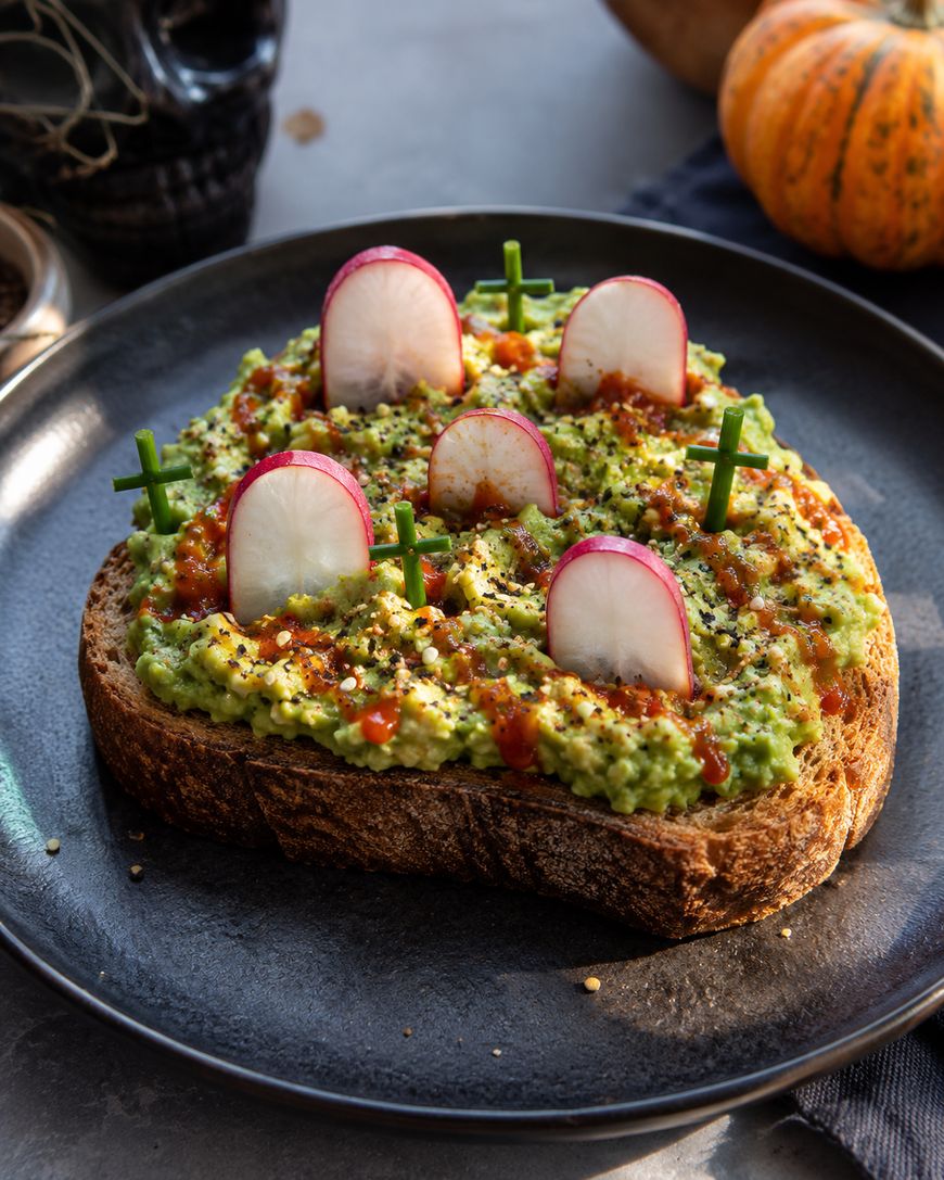 A slice of toast topped with guacamole, radish slices, and green garnish, arranged to look like a spooky graveyard scene with crosses, on a black plate.