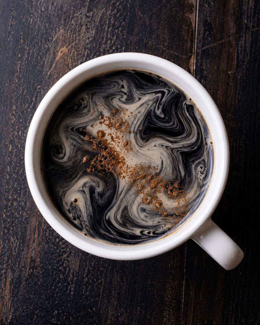 A white mug filled with black coffee topped with swirling cream and a sprinkle of cinnamon, placed on a dark wooden surface.