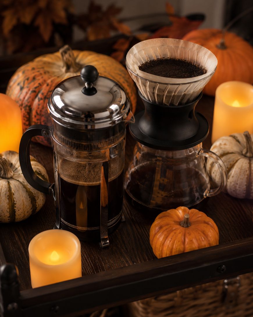 A French press and pour-over coffee maker sit on a table with lit candles and assorted pumpkins, creating a cozy autumn atmosphere.