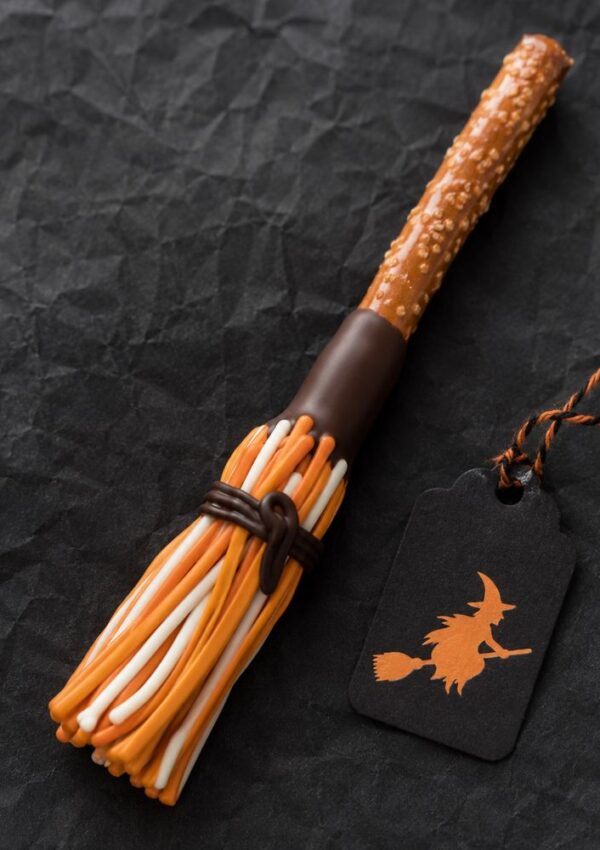 A broomstick-shaped Halloween treat made of a pretzel stick, candy strands, and chocolate sits next to a black tag with an orange witch silhouette—perfect for adults to enjoy or to include in Halloween goodie bags filled with festive treats.