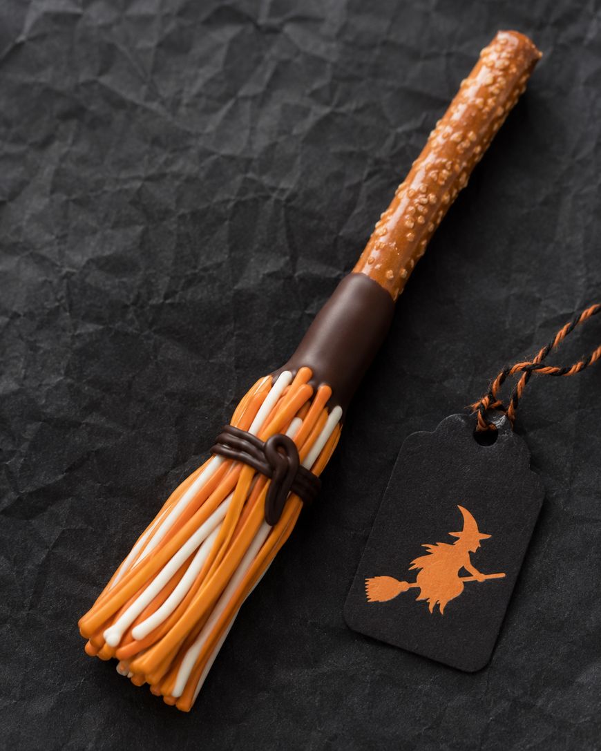 A broomstick-shaped Halloween treat made of a pretzel rod and orange, yellow, and white candy, tied with chocolate, next to a black tag featuring an orange silhouette of a witch on a broom.