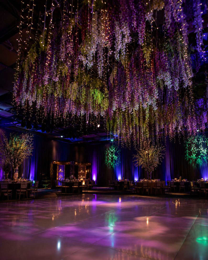 A ballroom with a glossy dance floor, hanging purple and green floral arrangements, string lights, and softly lit tables, creating an elegant, magical atmosphere.