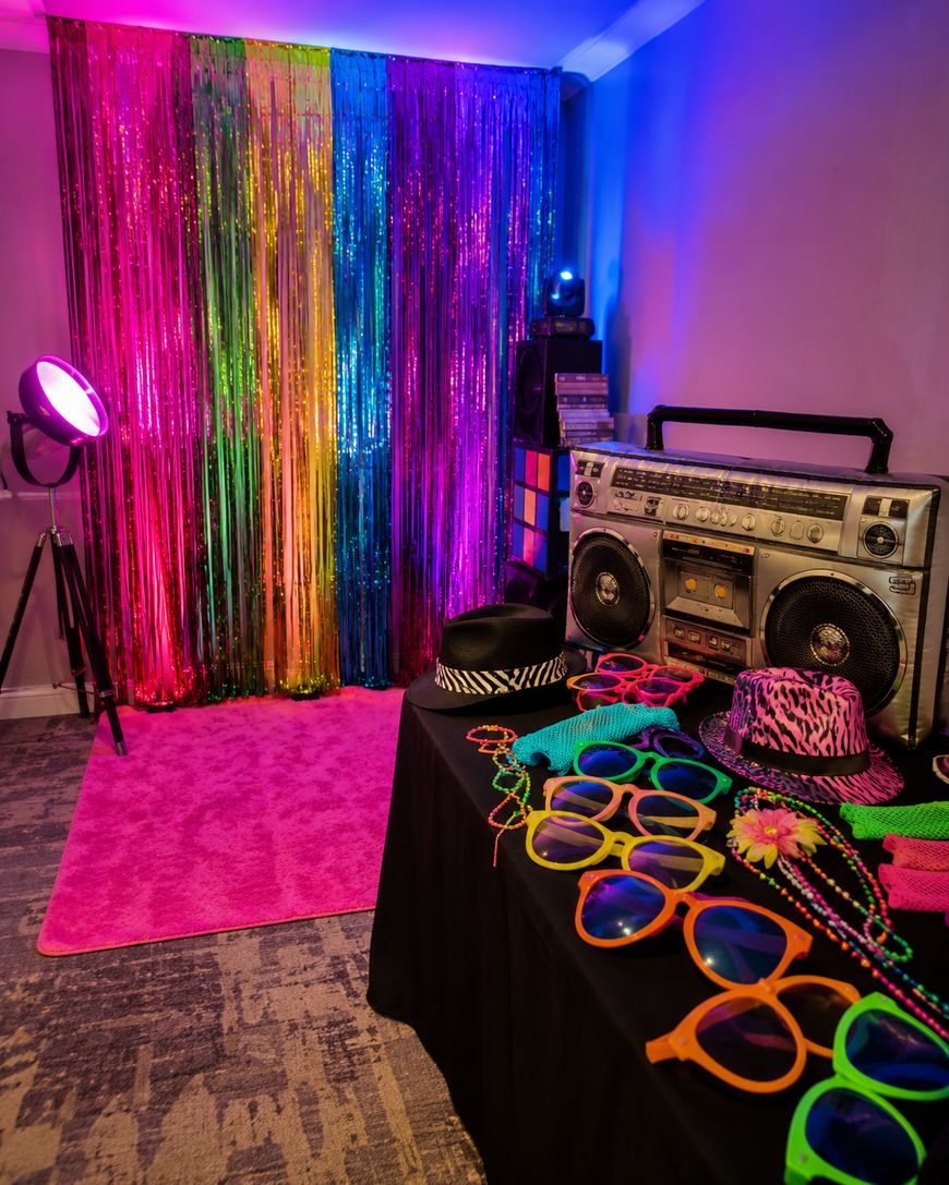 A colorful room with rainbow streamers, a pink rug, a spotlight, a retro boombox, and a table covered with neon hats, glasses, and accessories.