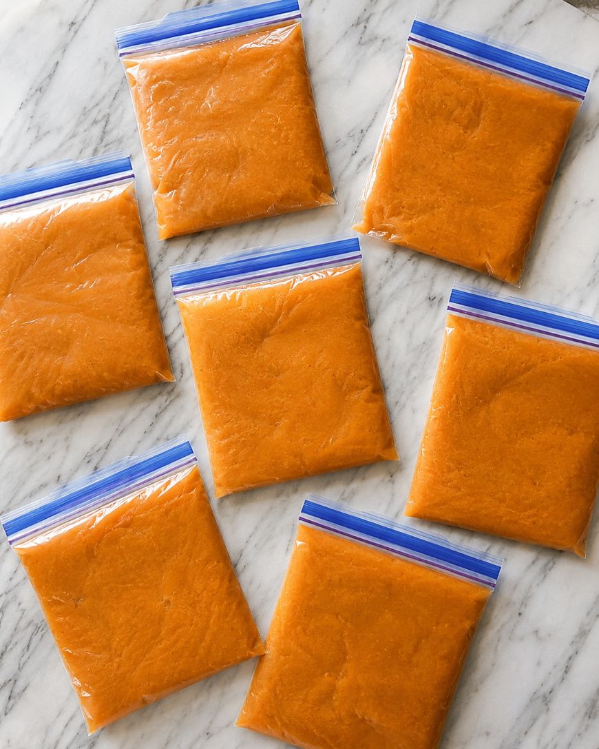 Seven zip-top bags filled with orange puree are arranged on a white marble surface. The bags are neatly sealed and spaced out, showing the contents clearly.