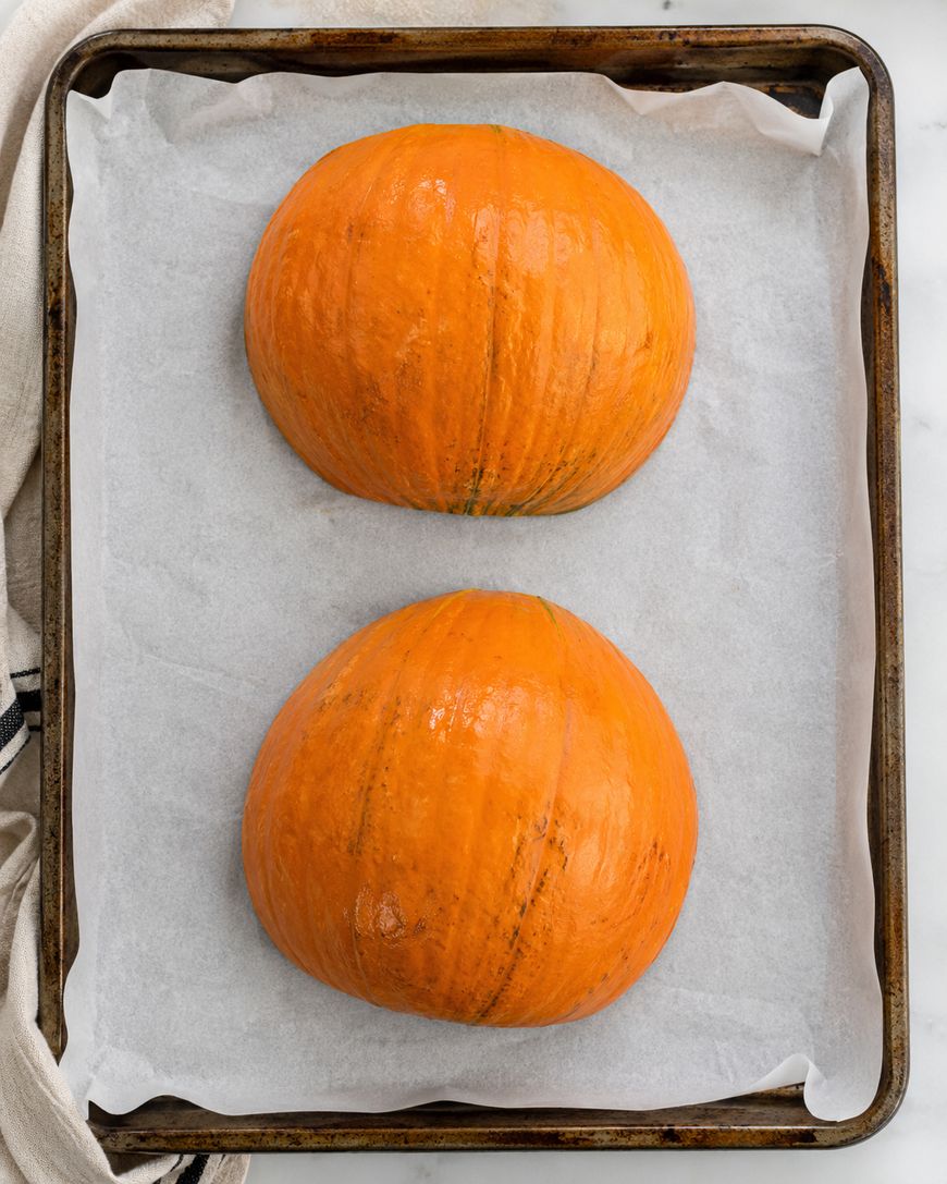 Two pumpkin halves, cut side down, on a parchment-lined baking sheet, ready to be roasted.