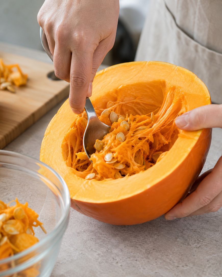 A person uses a spoon to scoop seeds and fibrous strands from a halved pumpkin, with a clear glass bowl nearby to collect the removed contents.