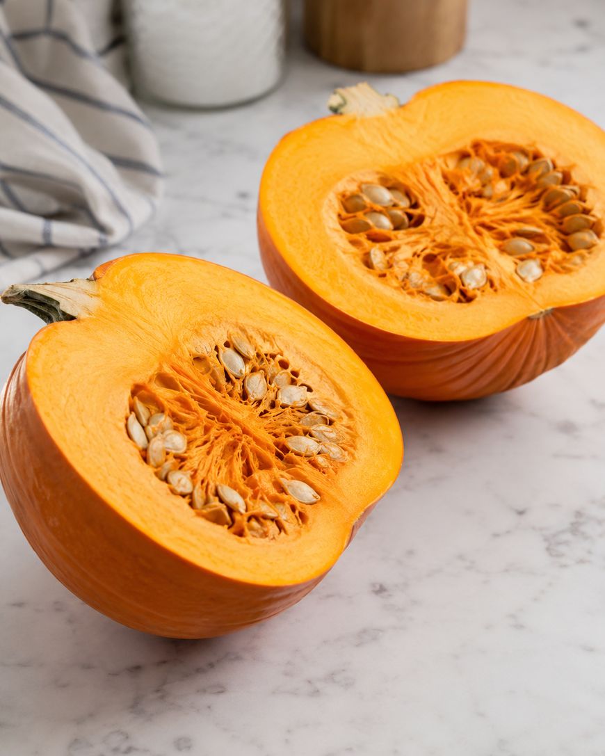 A pumpkin cut in half, showing its orange flesh and seeds, sits on a white marble countertop with a striped cloth and jars in the background.