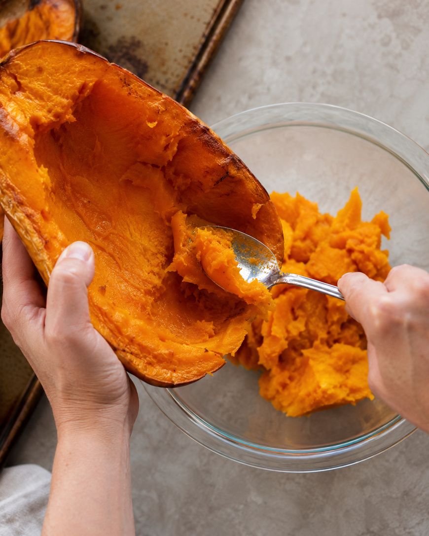A person scoops cooked butternut squash from the skin into a glass bowl using a spoon.