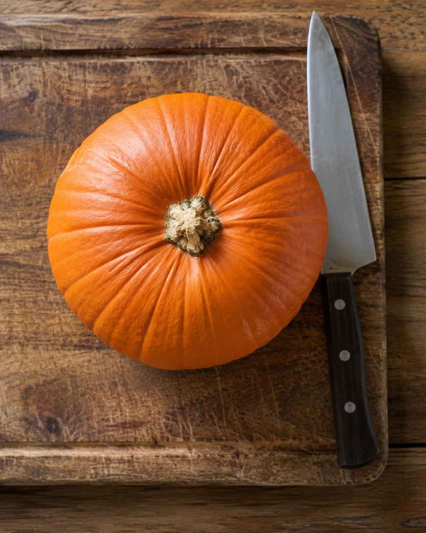 A whole orange pumpkin sits on a wooden cutting board next to a kitchen knife with a dark handle.