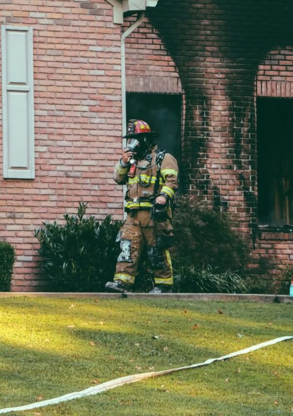 A firefighter in full gear stands outside a brick house with burned windows and smoke marks, holding a radio or mask, responding to a recent fire.