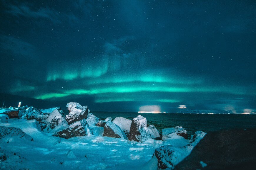 Snow-covered rocks by a cold sea under the night sky, illuminated by glowing green aurora borealis. City lights are faintly visible on the horizon in the distance.