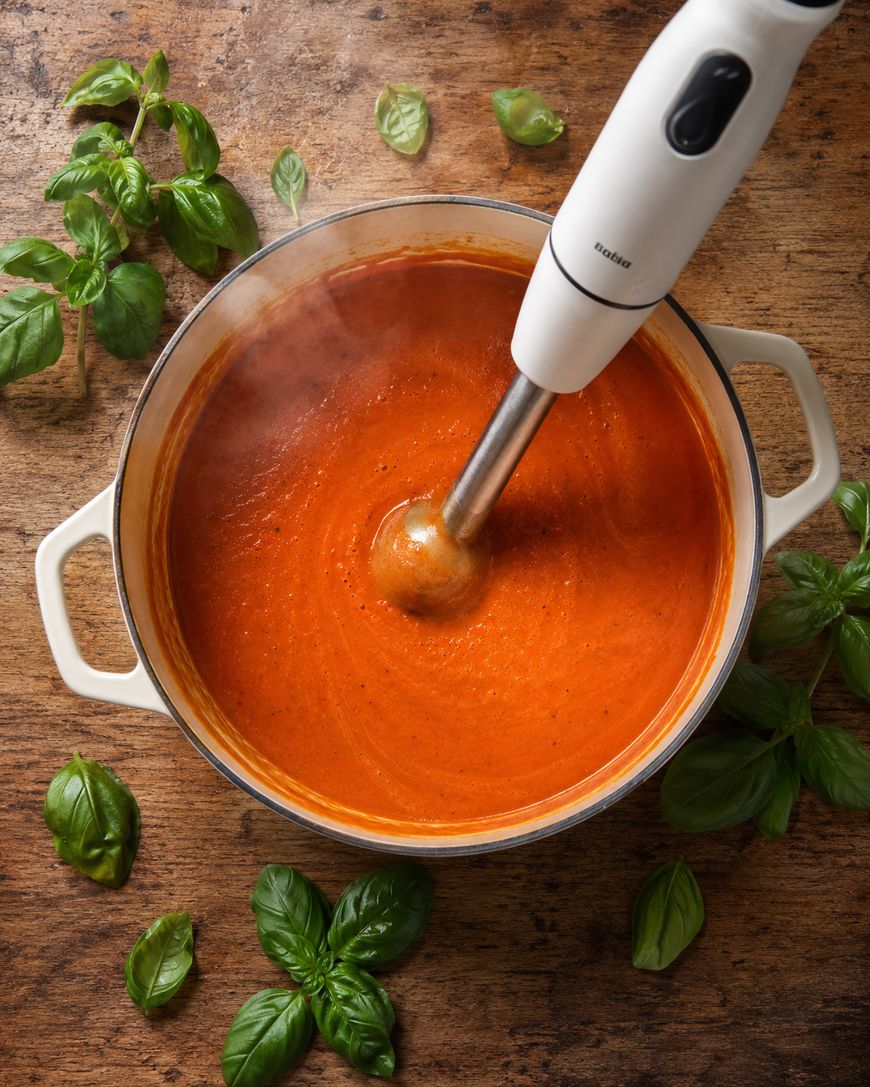 A white pot of tomato soup is being blended with an immersion blender on a wooden surface, surrounded by fresh basil leaves.