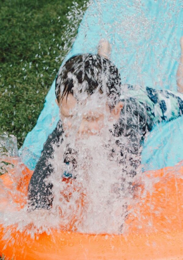 A child slides on an orange and blue slip-and-slide, splashing water everywhere, with grass visible on the side.