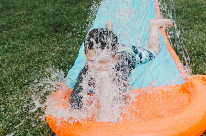 A child slides face-first down an orange and blue slip-and-slide, water splashing up and obscuring part of their face, on green grass.